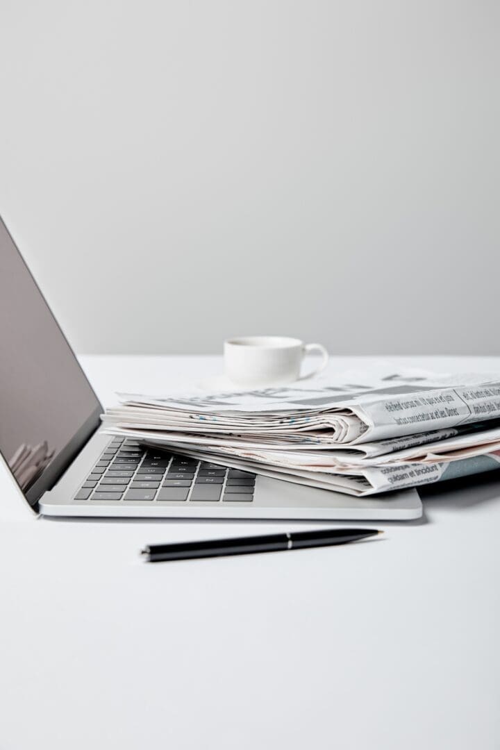 selective focus of laptop with blank screen near newspapers, pen and cup on grey